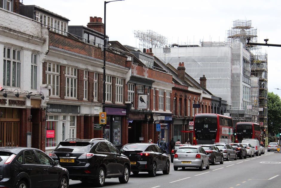 A street scene on Surbiton High Street showing a row of old brick and white-painted shops with large windows, some featuring signage for local businesses. Several cars are parked along the curb, inclu