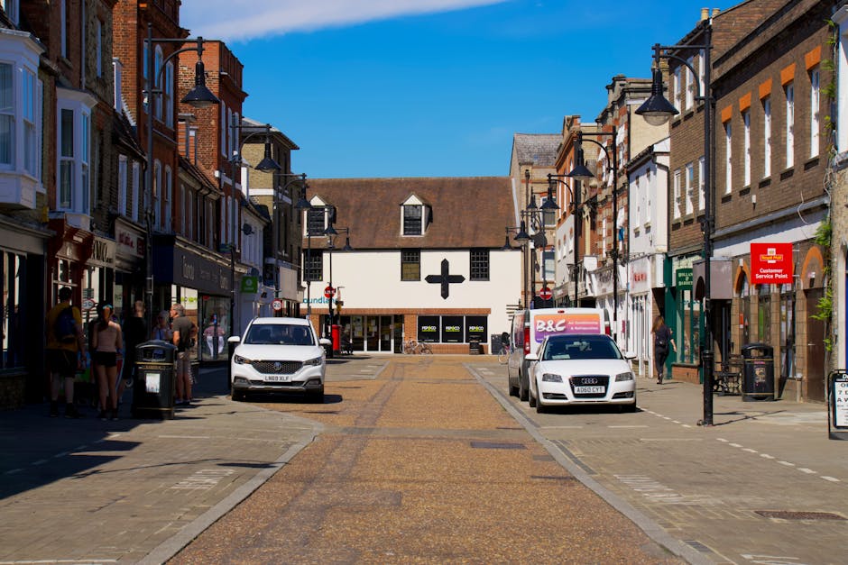 A street scene on Surbiton High Street showing a row of old brick and white-painted shops with large windows, some featuring signage for local businesses. Several cars are parked along the curb, inclu