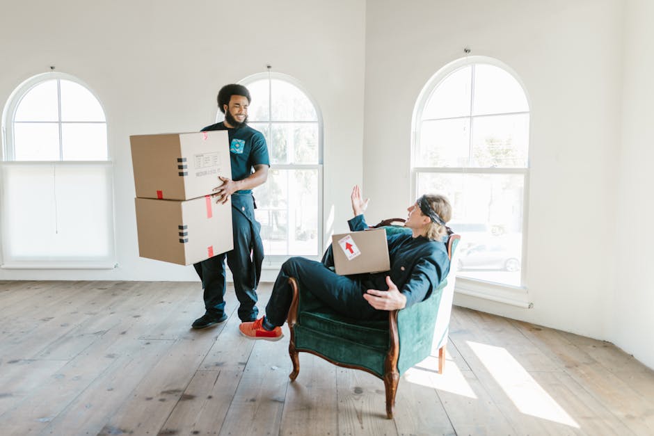 A man with dark hair and a beard, dressed in a service uniform, is carrying two large cardboard boxes wrapped with red and black tape, as part of the house removal process inside a bright, spacious ro