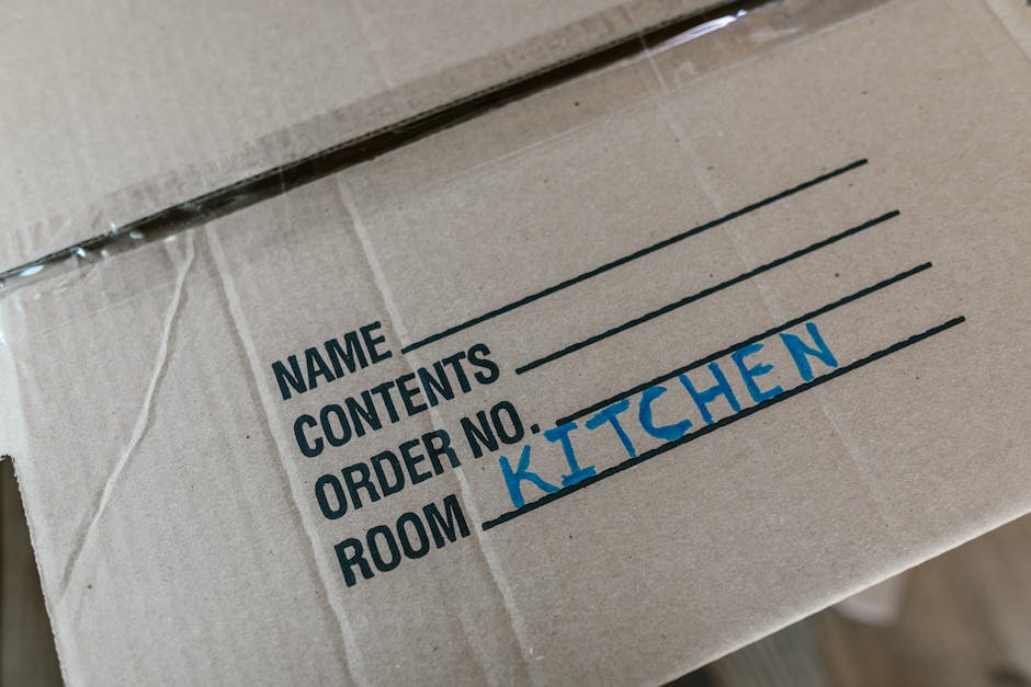 A man with dark hair and a beard, dressed in a service uniform, is carrying two large cardboard boxes wrapped with red and black tape, as part of the house removal process inside a bright, spacious ro