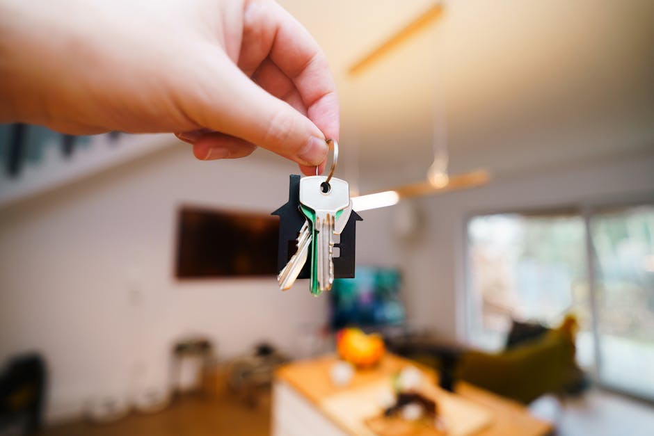 A close-up image of a hand holding a keyring with several keys, including a silver key and a green plastic key, suspended in the air inside a home. The background reveals a living room with a large wi